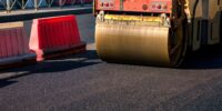 Workers are engaged in asphalt paving on a city road, utilizing heavy machinery to ensure a smooth surface. Bright sun illuminates the area, revealing safety barriers and ongoing construction efforts.