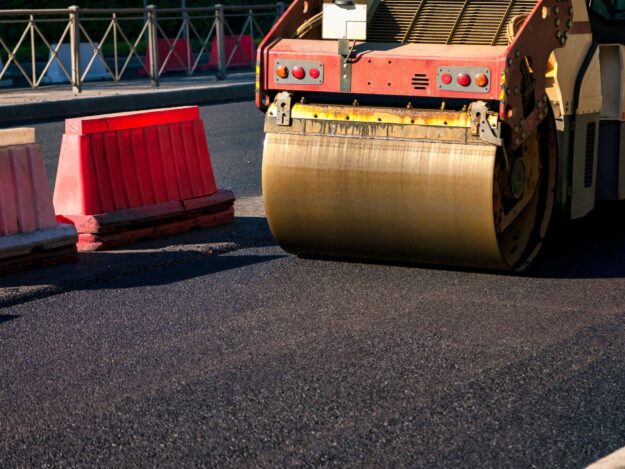 Heavy Roller Compacts Freshly Paved Asphalt on Urban Road Construction Site Workers are engaged in asphalt paving on a city road, utilizing heavy machinery to ensure a smooth surface. Bright sun illuminates the area, revealing safety barriers and ongoing construction efforts.